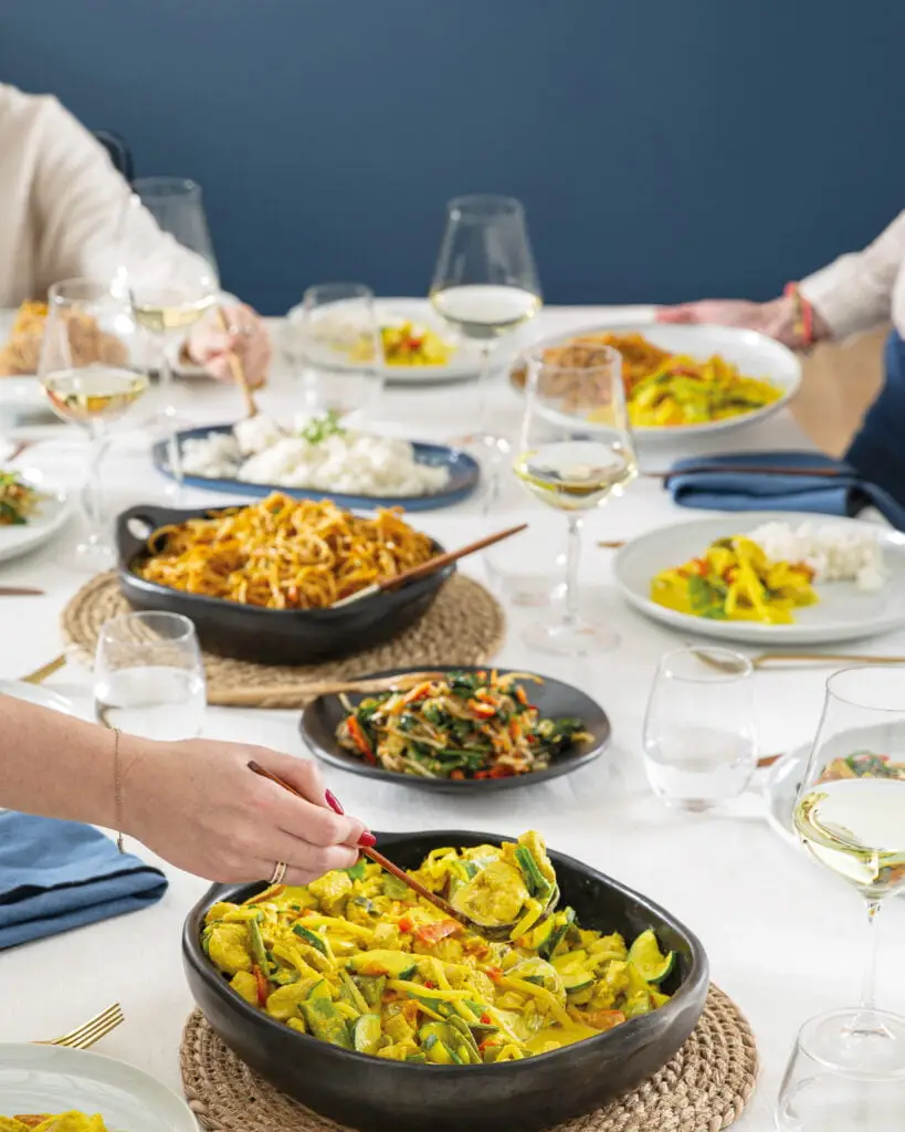 La photo montre une table avec plusieurs plats de cuisine asiatique, des baguettes et des verres de vin blanc. Une personne se sert dans les grands plats avec une cuillère en bois pour servir les invités.